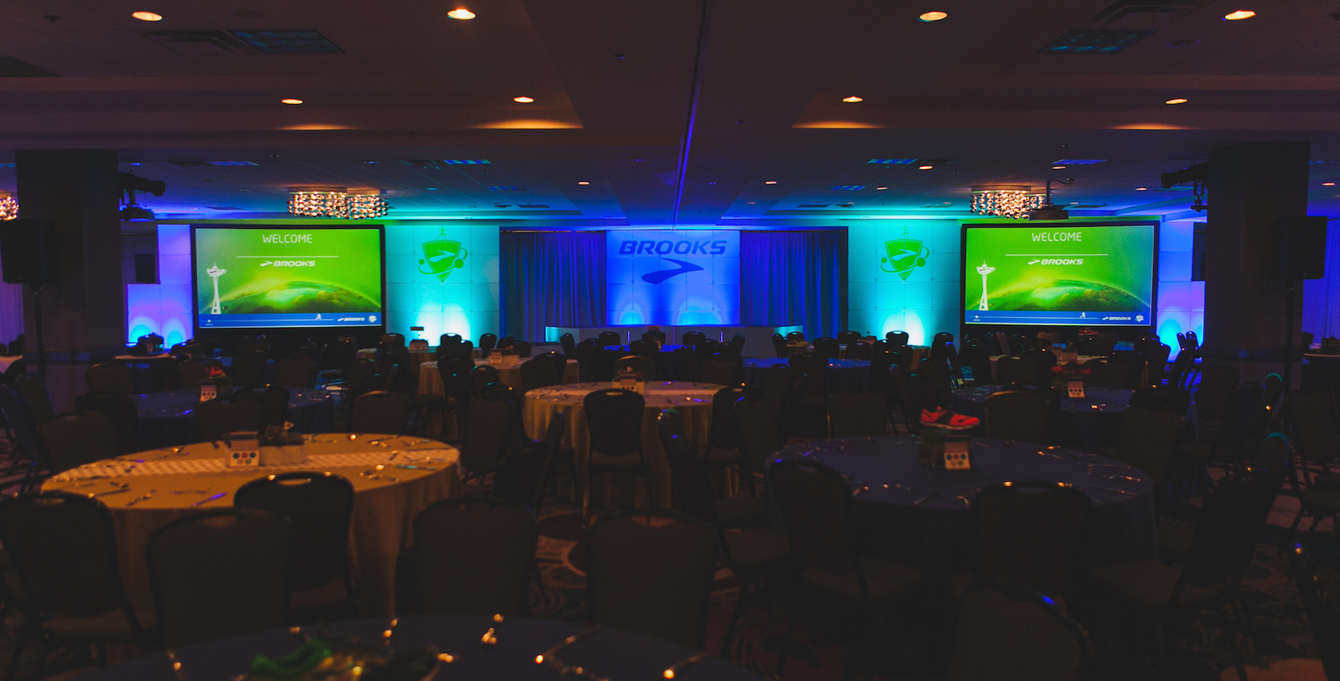 Two big screens dispalying logos are on either side of a stage for a corporate event. There is blue and green lighting and an impressive atmosphere.